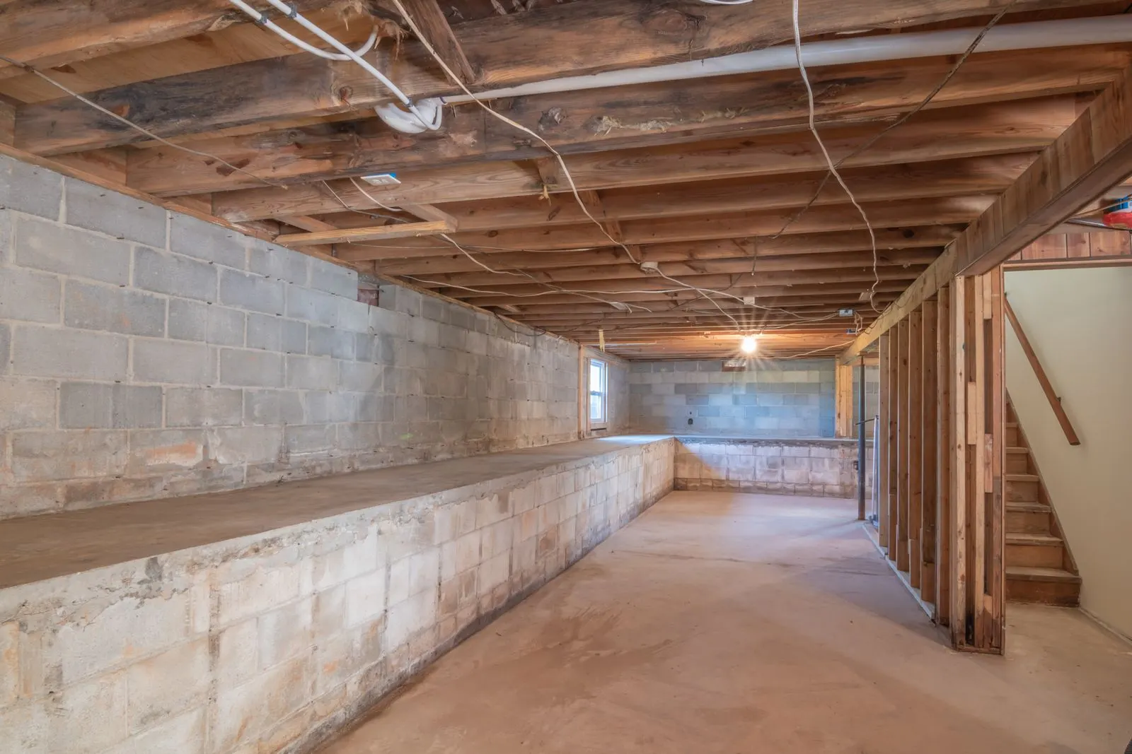 Interior of an unfinished basement showing a concrete block foundation wall, poured floor and exposed wood floor joists overhead