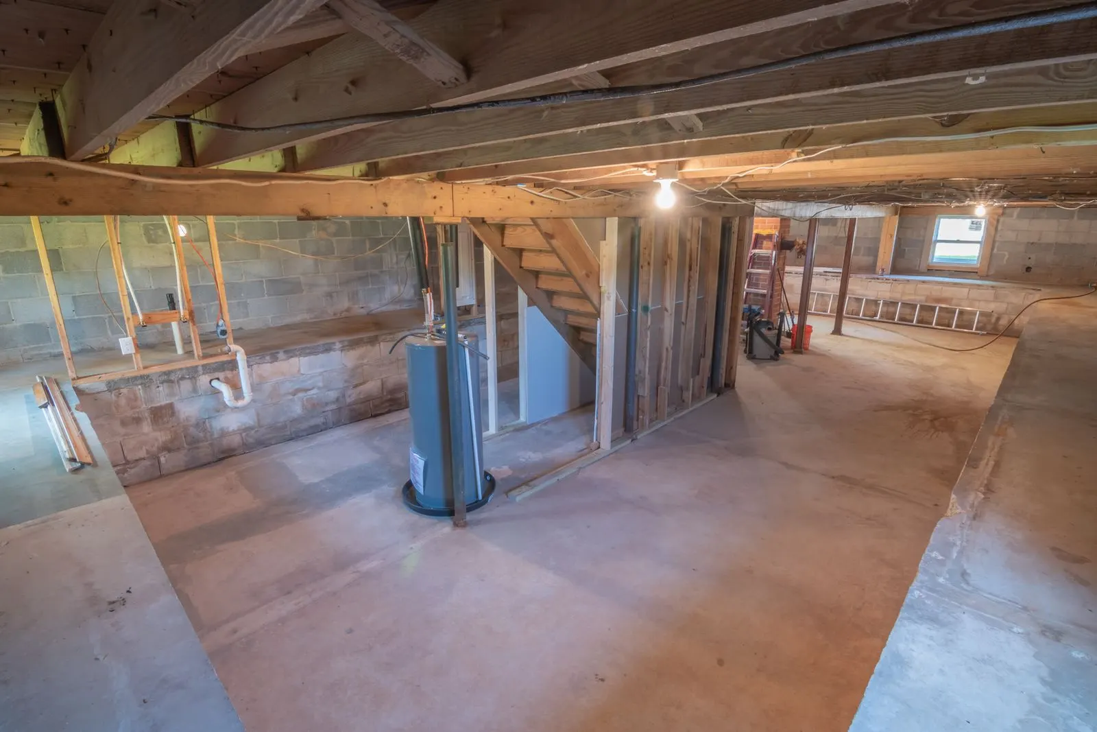 Unfinished basement with exposed wood floor joists, concrete block foundation walls, a water heater and overhead plumbing runs