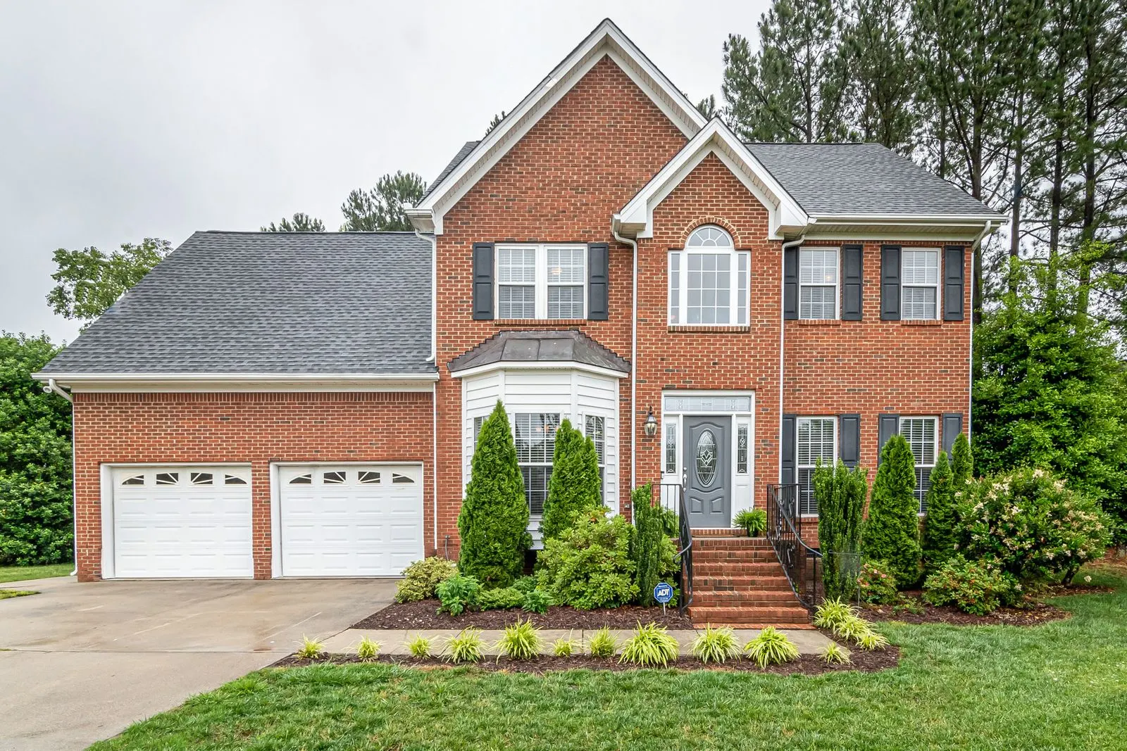 Two-storey red brick family home with twin white garage doors, dark shutters, foundation plantings and pine trees behind