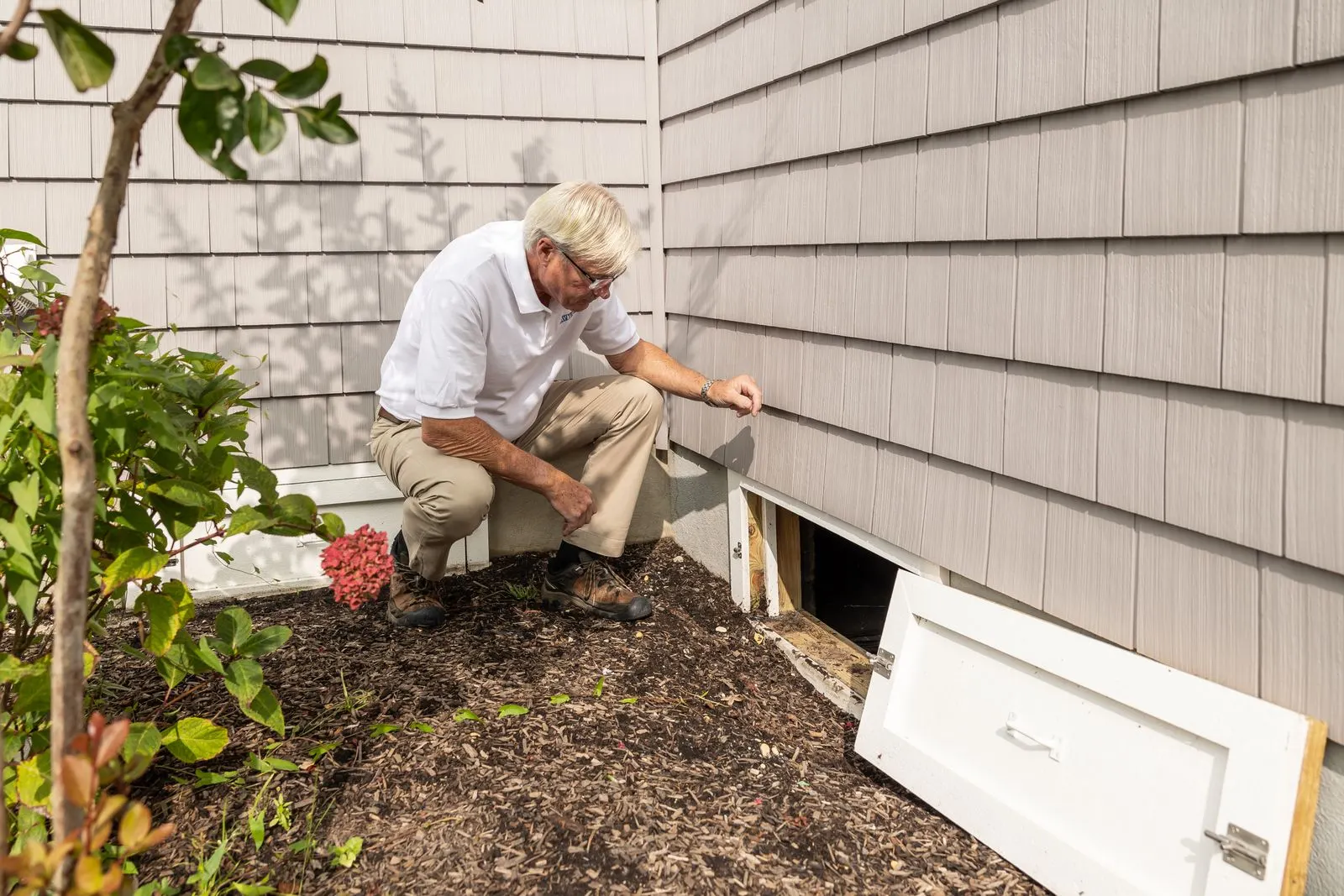 A person kneeling beside an open crawl space access door on the exterior of a shingle-sided house, with the removed access panel leaning against the wall
