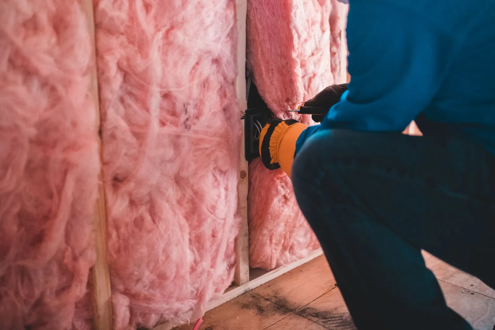 Worker in gloves fitting pink fiberglass batt insulation between wood framing members