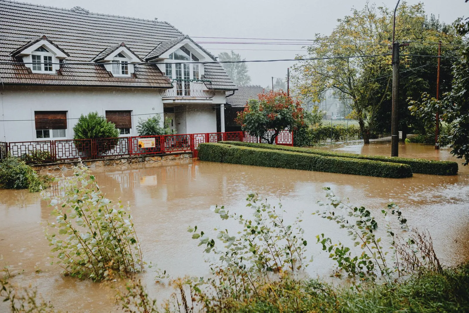 A suburban house surrounded by standing brown floodwater after heavy rain, with water reaching the garden wall and hedge