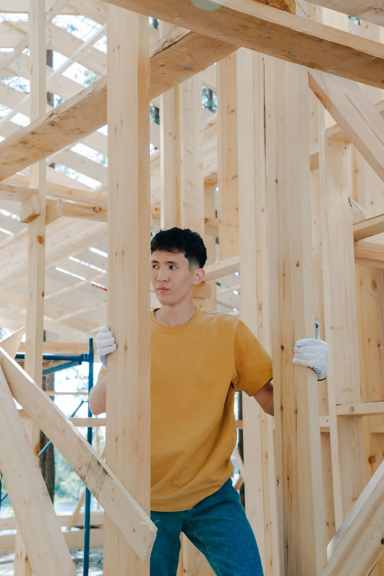 Construction worker in gloves steadying a vertical wood framing member inside a partially built timber-framed structure